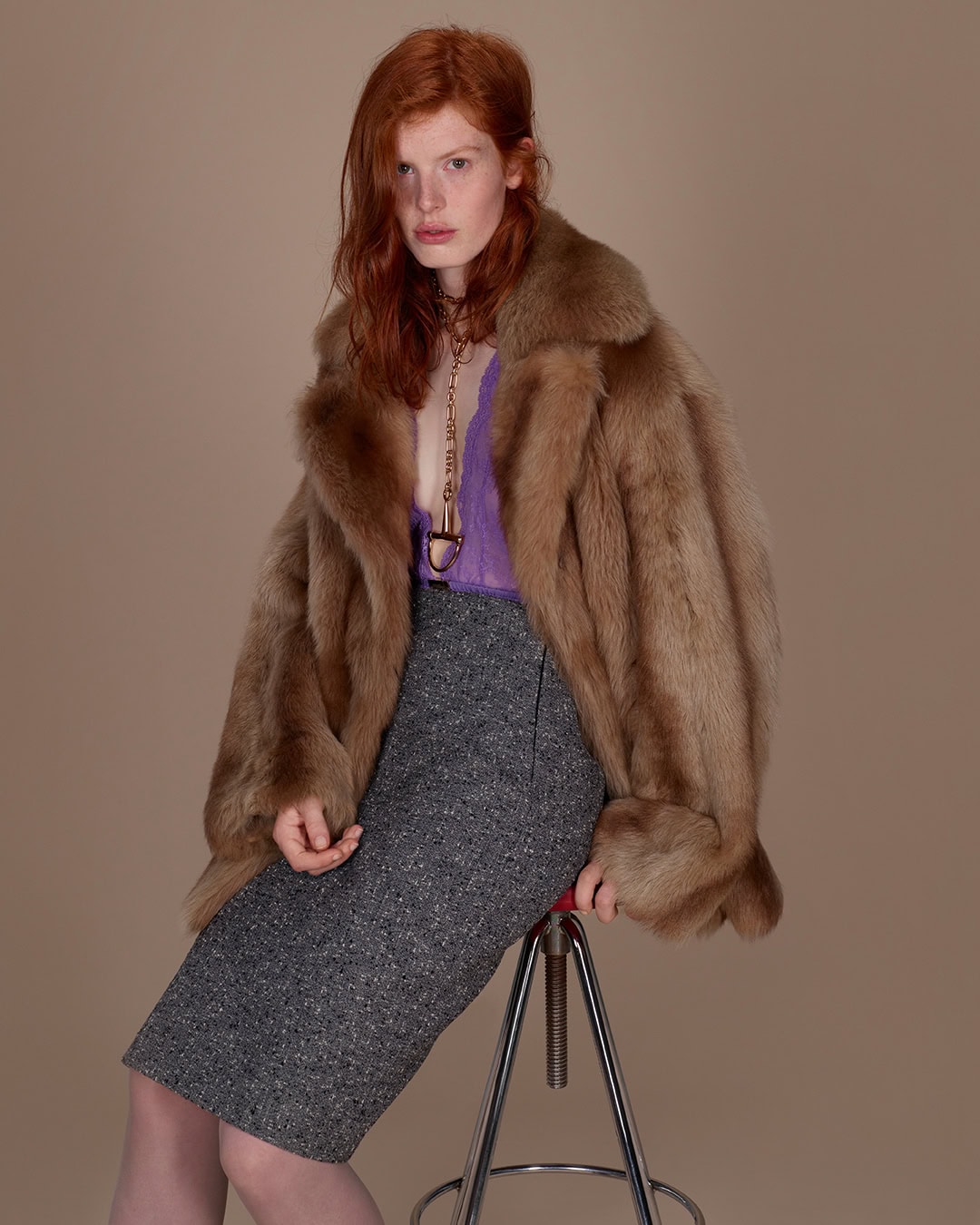 Red-haired model wearing a fur coat and grey skirt seated on a stool for Gucci
