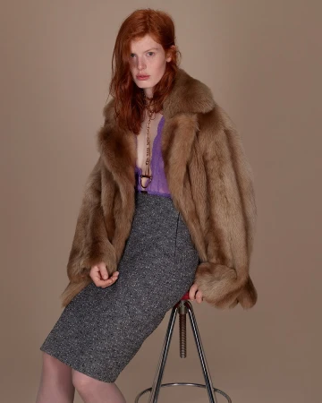 Red-haired model wearing a fur coat and grey skirt seated on a stool for Gucci