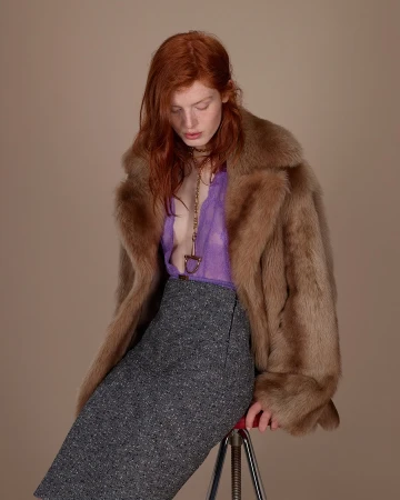 Red-haired model wearing a fur coat, purple lace top, and tweed skirt seated on a stool against a neutral backdrop.