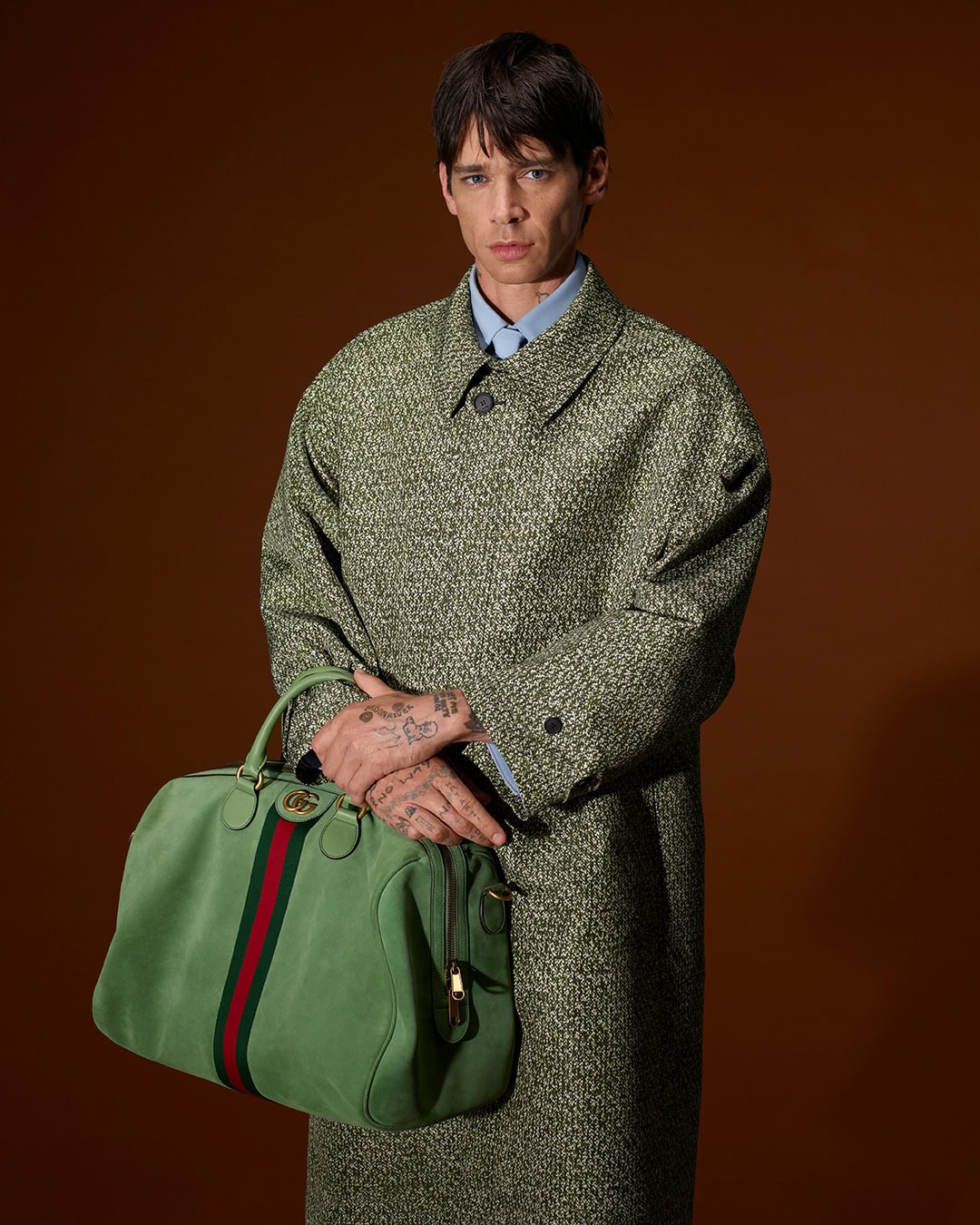 Man wearing a textured green coat holding a light green duffle bag against a dark brown background