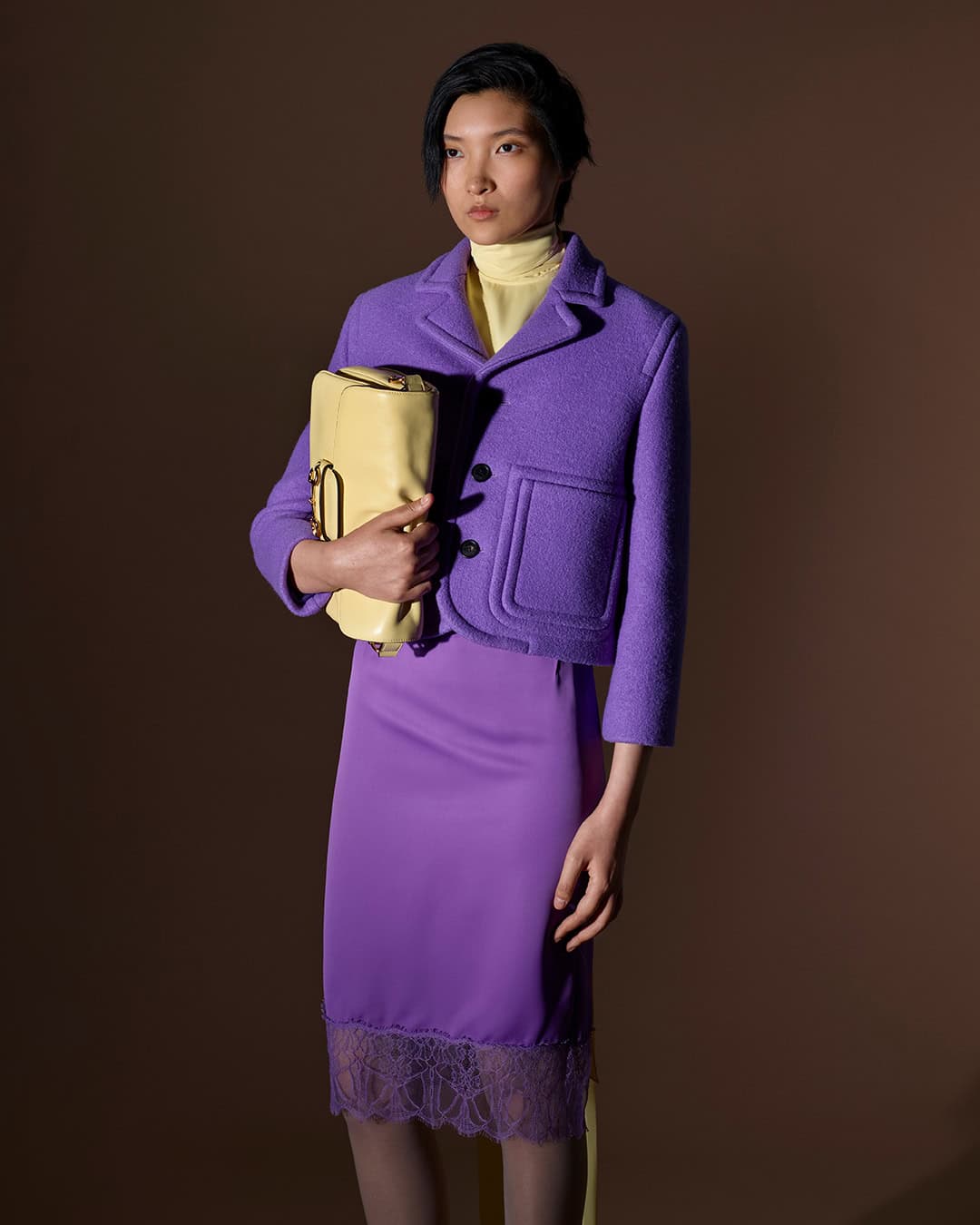 Model wearing a purple jacket and skirt holding a yellow handbag against a solid brown background