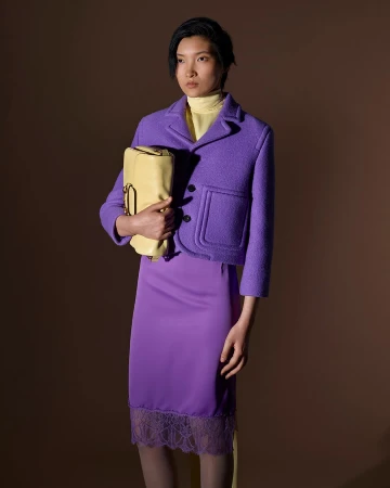 Model wearing a purple jacket and skirt holding a yellow handbag against a solid brown background
