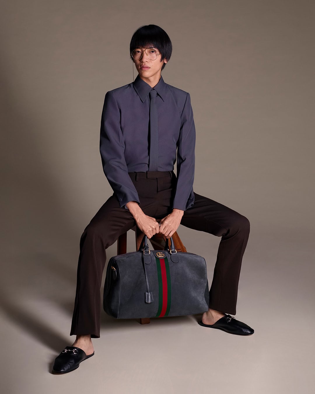 Man wearing slate grey button-down shirt and dark trousers holding a duffel bag while seated on a wooden stool in a studio.