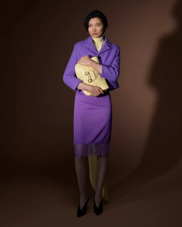 Model wearing a purple jacket and skirt holding a yellow handbag against a brown studio backdrop.