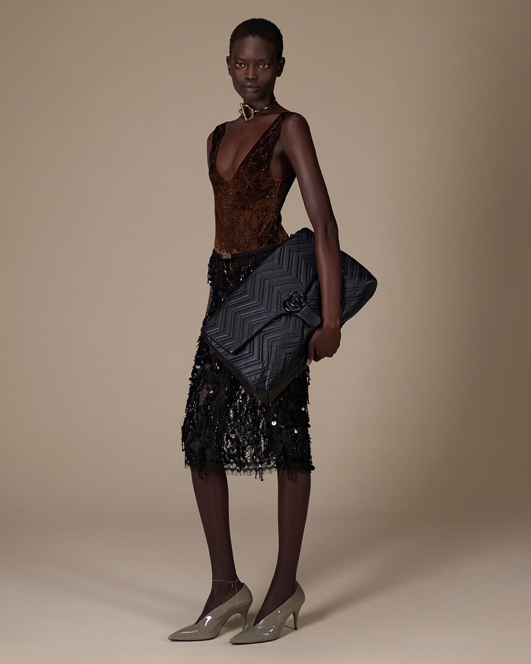 Model standing against a neutral gradient background wearing a dark top and skirt while holding a large black quilted handbag