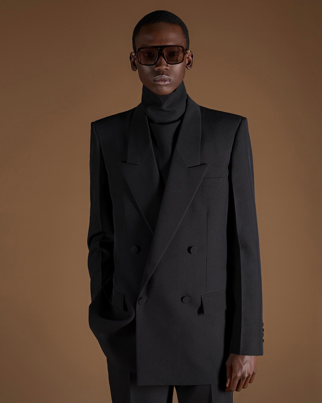 Model wearing a structured black double-breasted blazer and turtleneck against a warm brown backdrop for Gucci.