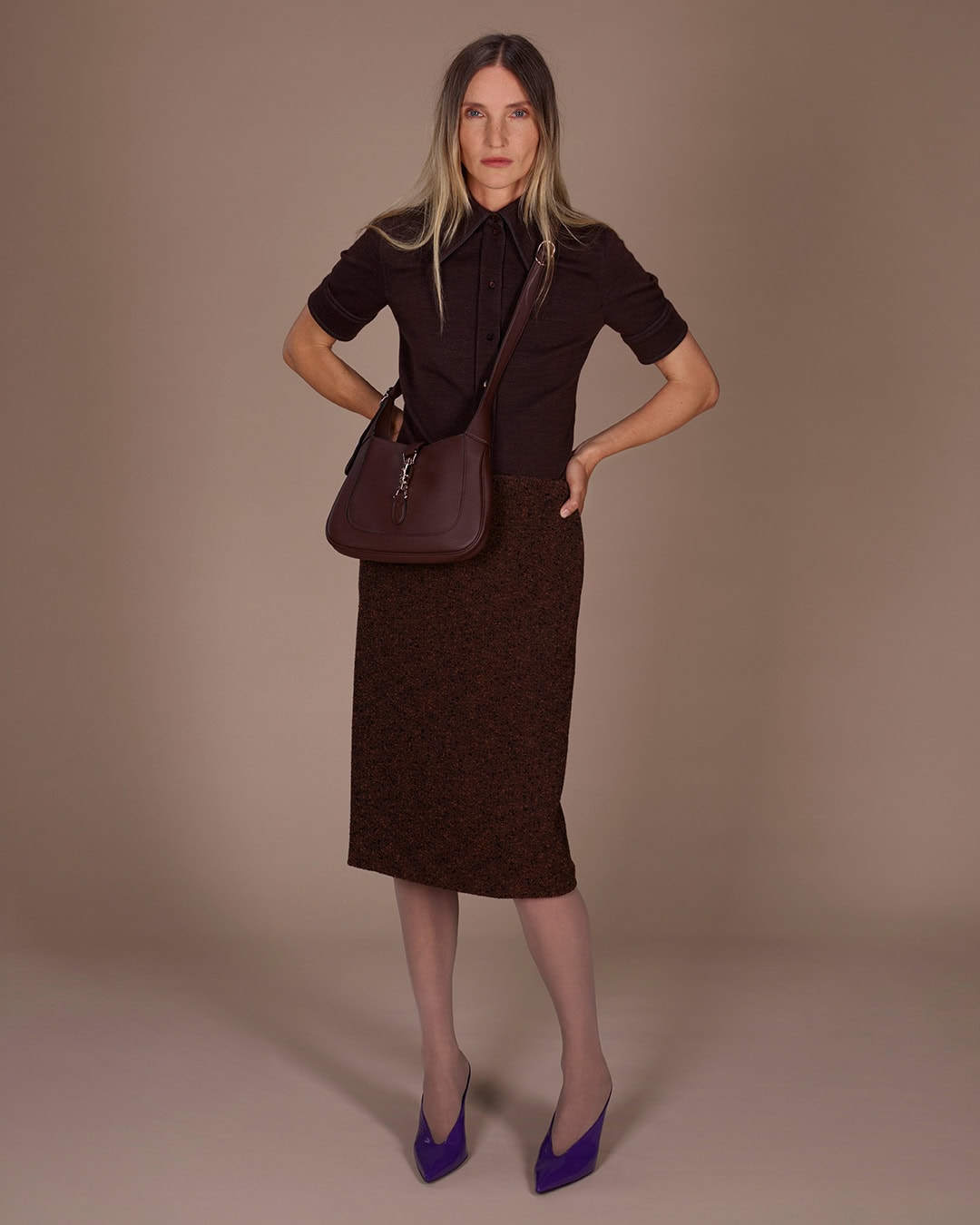 Model wearing dark brown polo shirt and textured skirt holding a shoulder bag against a neutral studio seamless background.