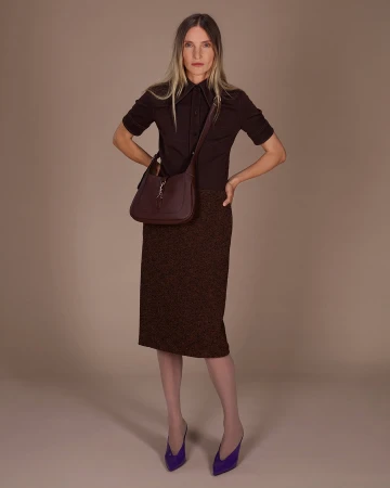Model wearing dark brown polo shirt and textured skirt holding a shoulder bag against a neutral studio seamless background.