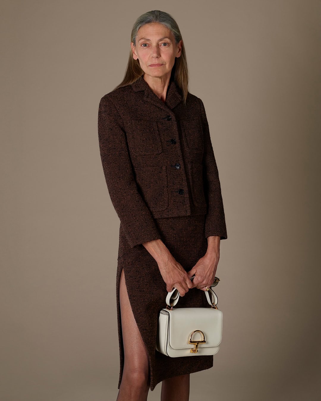 Elderly woman in a brown tweed jacket and skirt holding a white handbag against a neutral studio background