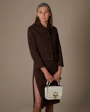 Elderly woman in a brown tweed jacket and skirt holding a white handbag against a neutral studio background