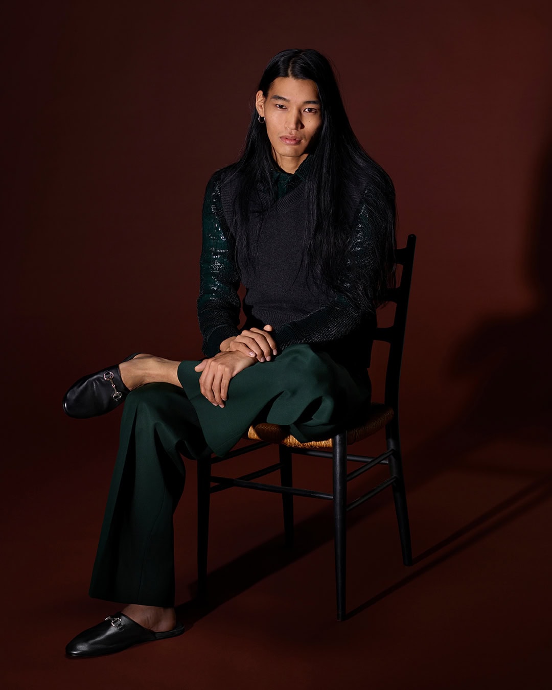 Man with long dark hair sitting on a dark wooden chair against a brown backdrop wearing a dark top and green trousers.