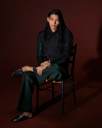 Man with long dark hair sitting on a dark wooden chair against a brown backdrop wearing a dark top and green trousers.
