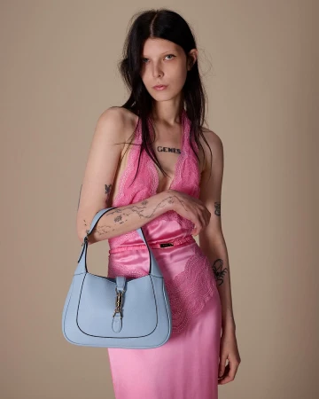 Model in a pink lace dress holding a light blue leather shoulder bag against a neutral background