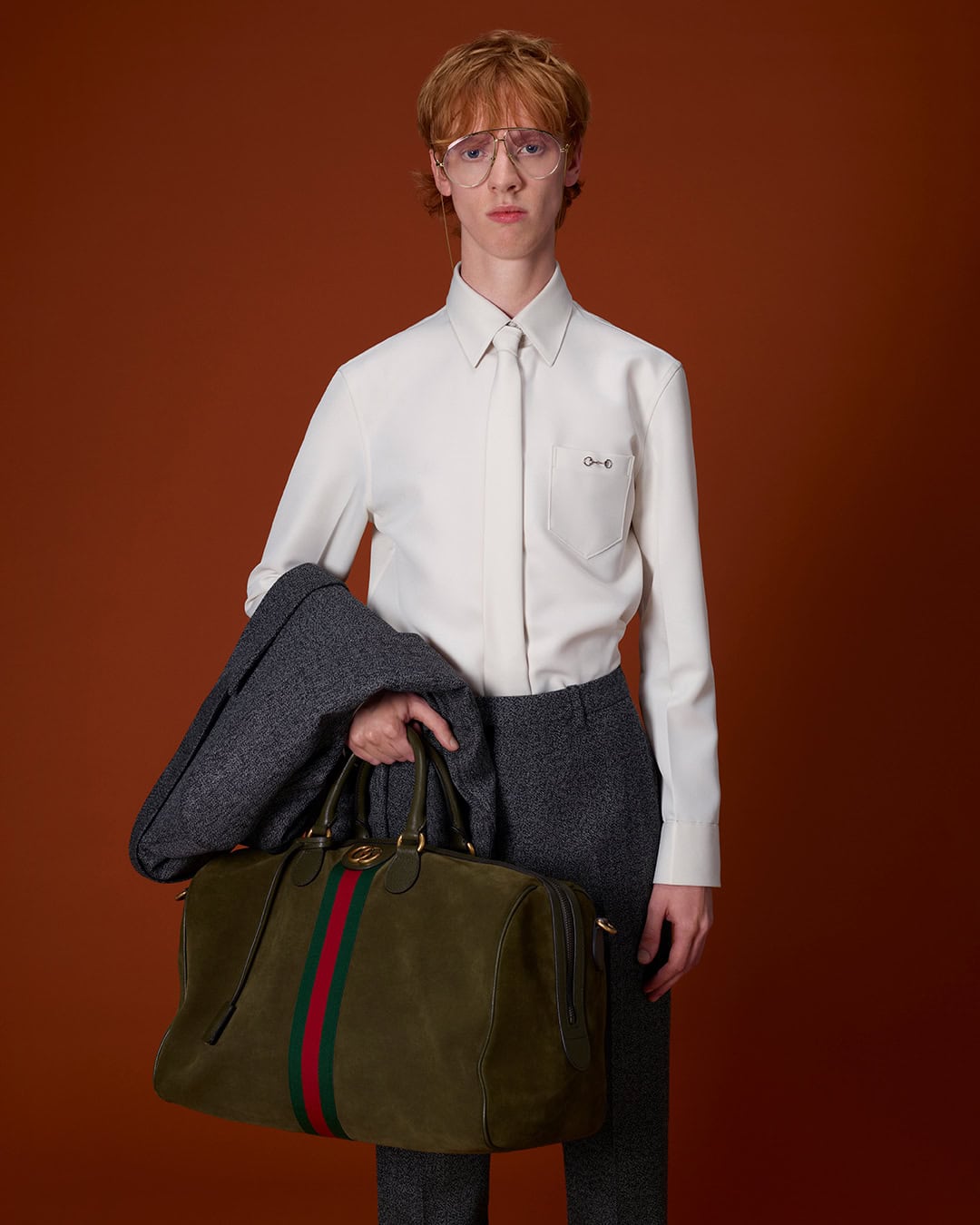 Model in white shirt and grey trousers holding an olive duffel bag against a solid rust-brown background for Gucci