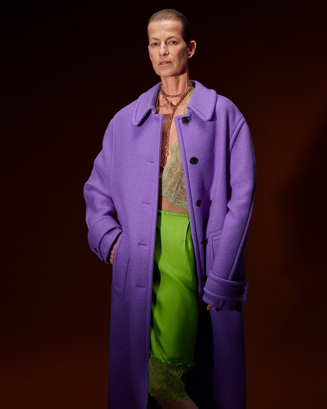 Model wearing a long purple coat over a green skirt against a dark background for the Gucci Fall 2025 campaign.