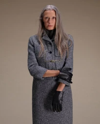 Elderly woman with long grey hair wearing a grey tweed suit and black leather gloves against a neutral background.