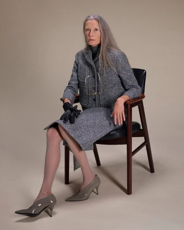 Elderly woman with long grey hair wearing a grey tweed skirt suit and heels, seated in an armchair against a neutral background.