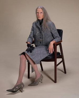 Elderly woman with long grey hair wearing a grey tweed skirt suit and heels, seated in an armchair against a neutral background.