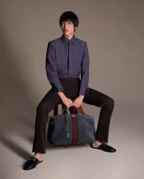 Man wearing slate grey button-down shirt and dark trousers holding a duffel bag while seated on a wooden stool in a studio.