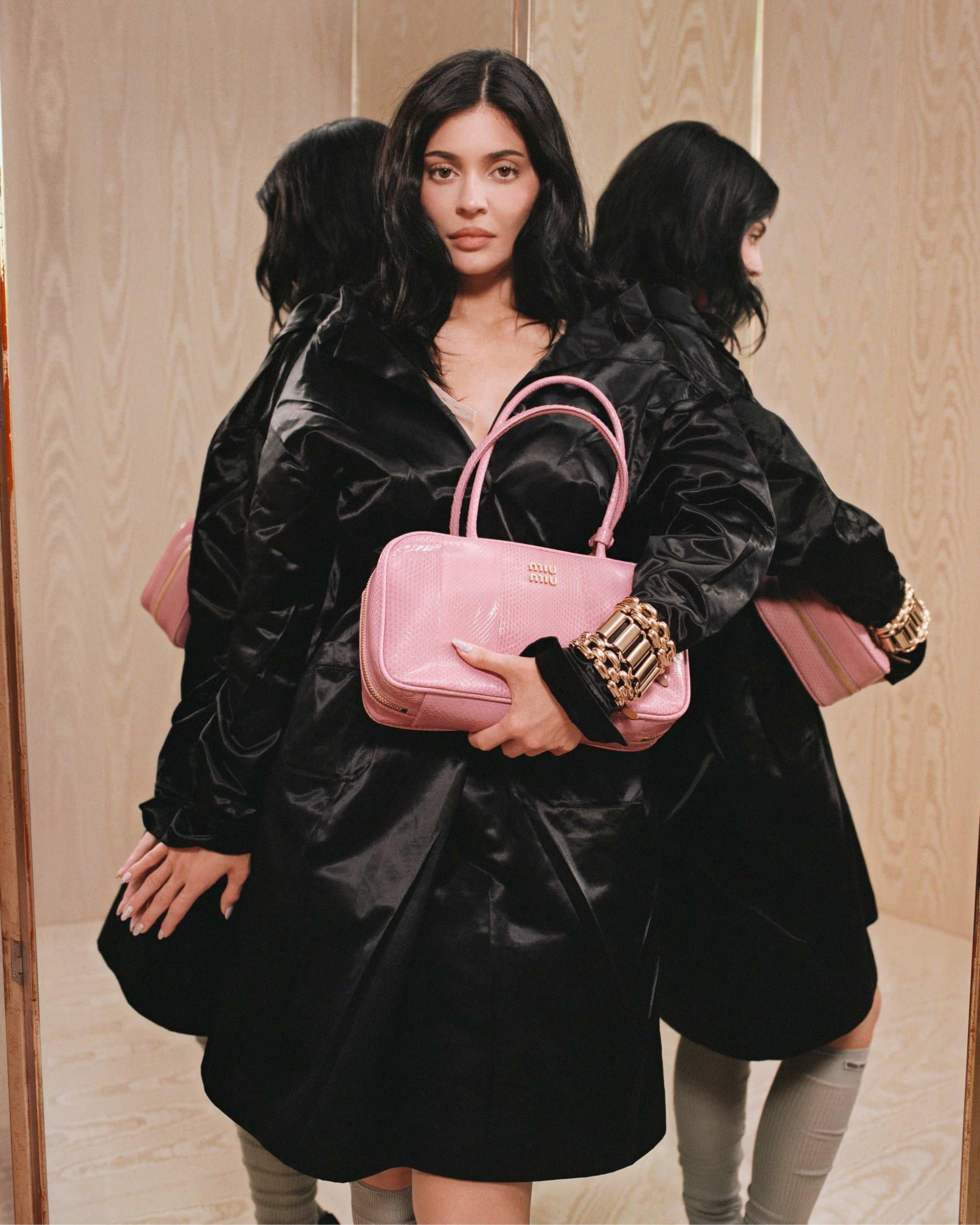 Kylie Jenner holding a pink handbag wearing a black coat against a wood paneled background
