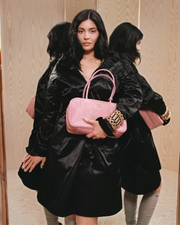 Kylie Jenner holding a pink handbag wearing a black coat against a wood paneled background