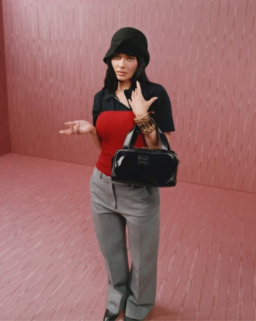 Kylie Jenner wearing a black hat, red top, grey trousers, and holding a black handbag against a pink textured wall.