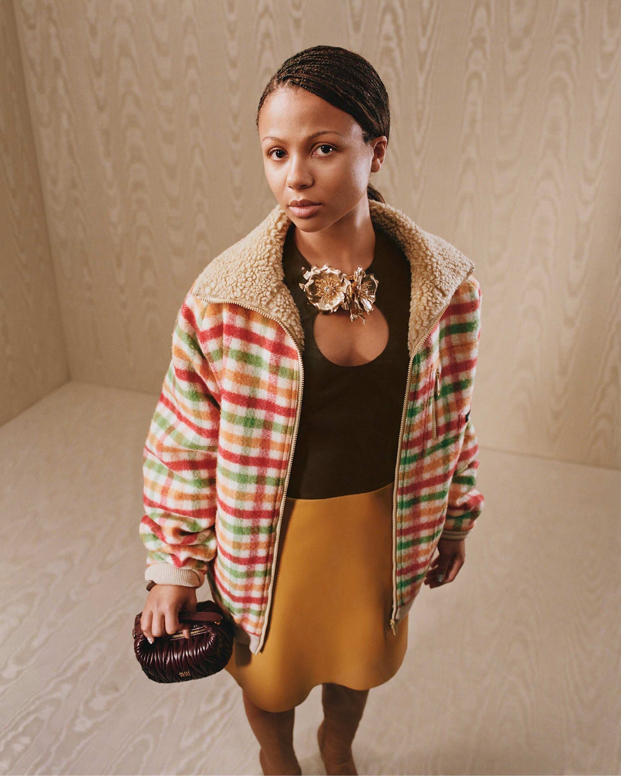 Model wearing a plaid sherpa jacket and yellow skirt holding a small dark handbag in front of a wood paneled wall