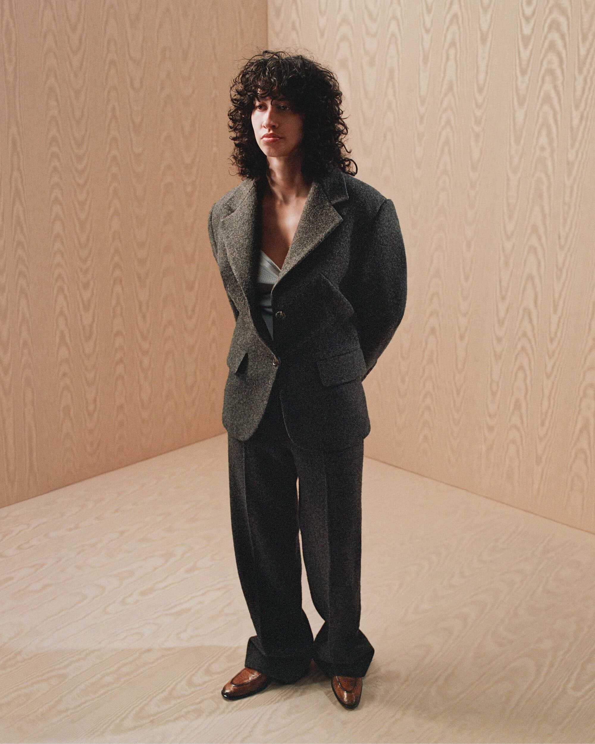 Model with dark curly hair wearing a dark grey suit standing in a corner with light wood paneled walls