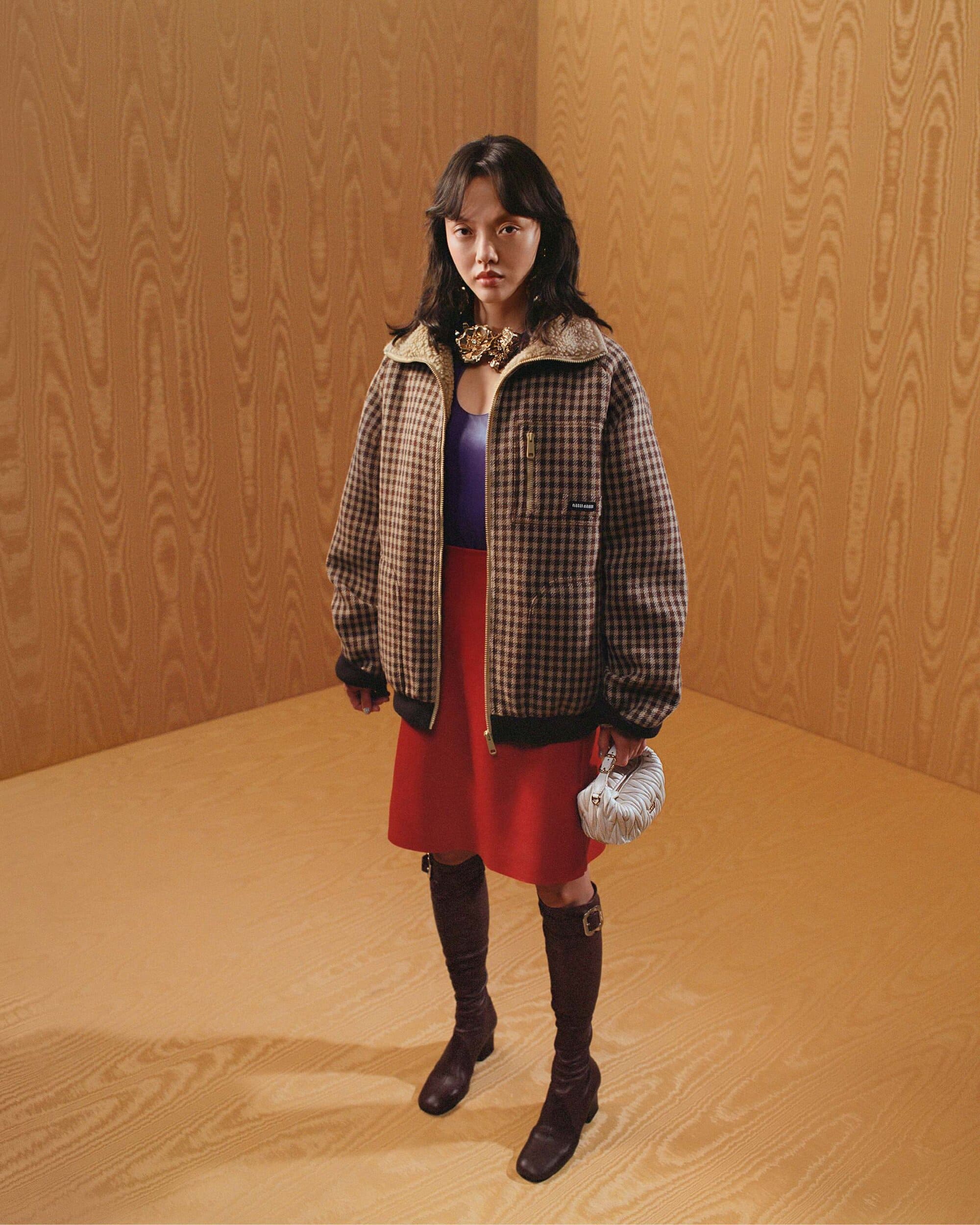 Model standing in front of light wood paneled walls wearing a brown patterned jacket red skirt and tall boots