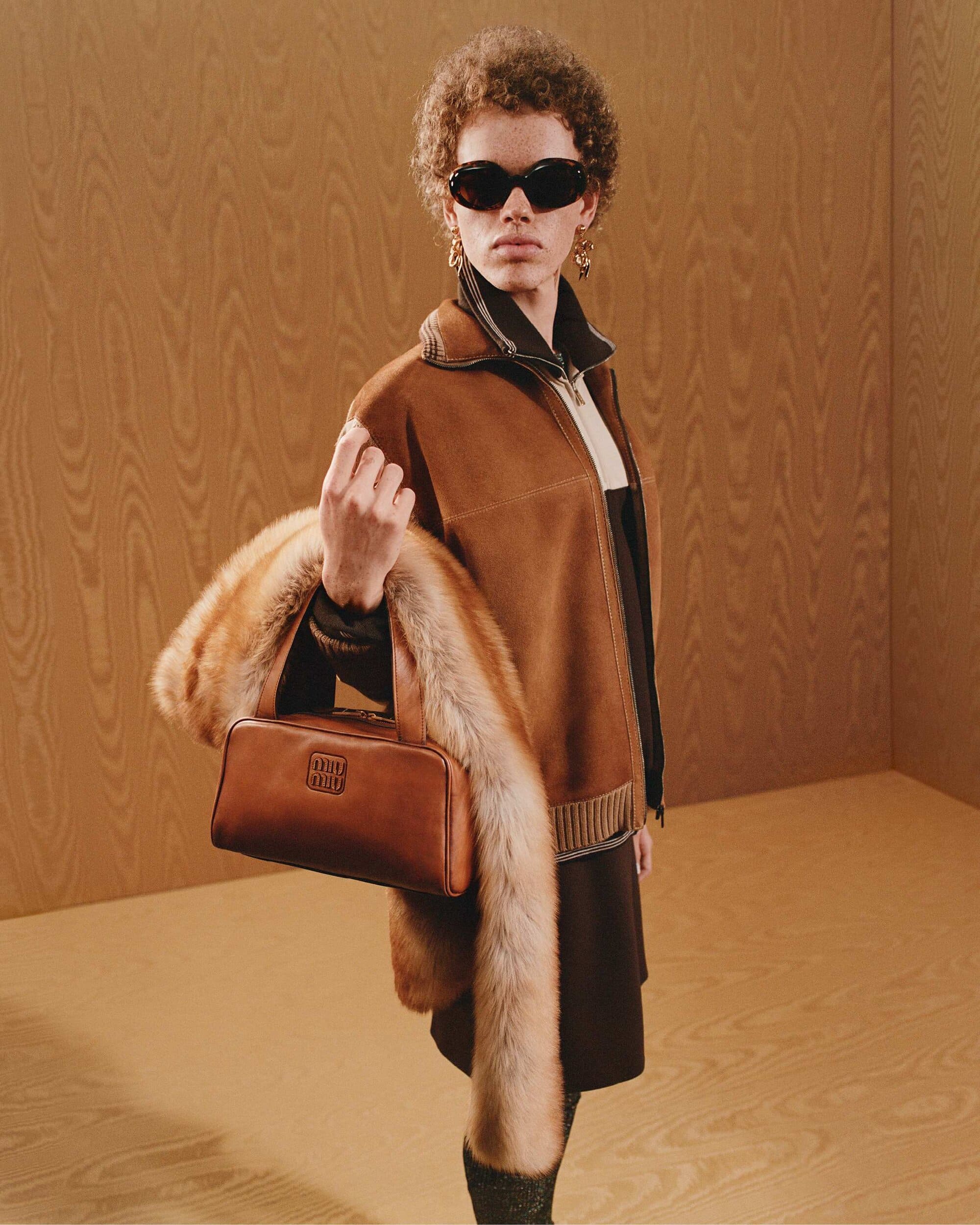 Model wearing dark sunglasses and a brown jacket holding a leather handbag and fur stole against wood-paneled walls.