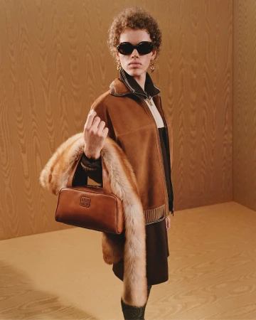 Model wearing dark sunglasses and a brown jacket holding a leather handbag and fur stole against wood-paneled walls.