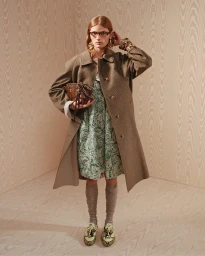 Model wearing a brown coat and patterned dress holding a handbag against a wood panel background