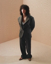 Model with dark curly hair wearing a dark grey suit standing in a corner with light wood paneled walls