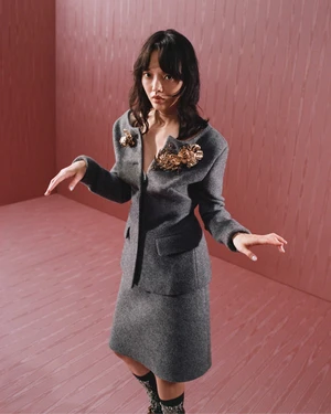 Model wearing a grey wool suit with ornate gold brooches in a pink paneled room for Miu Miu Fall 2025 ad campaign.