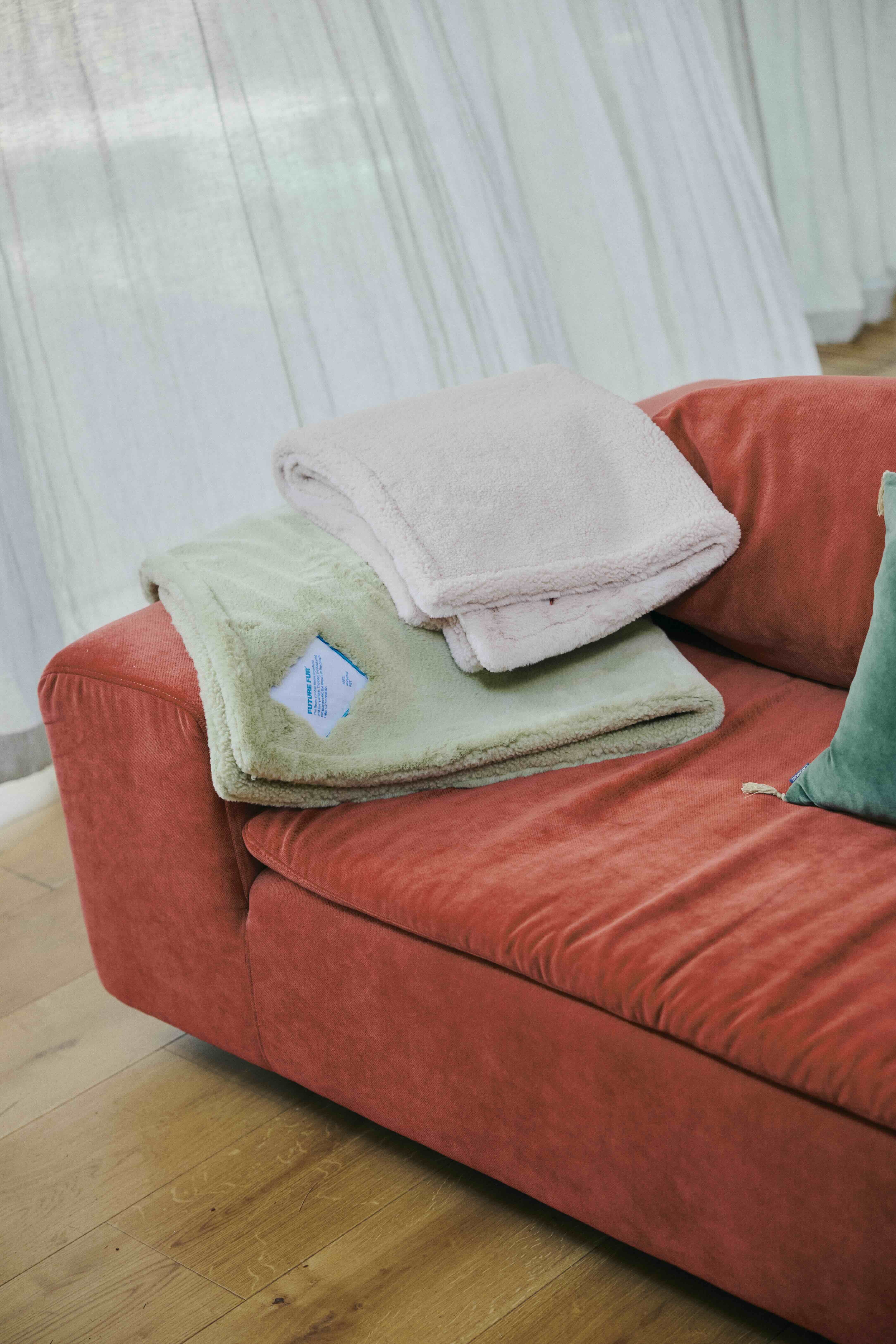 Folded sage green and cream faux fur blankets resting on a vibrant red sofa with a small teal pillow