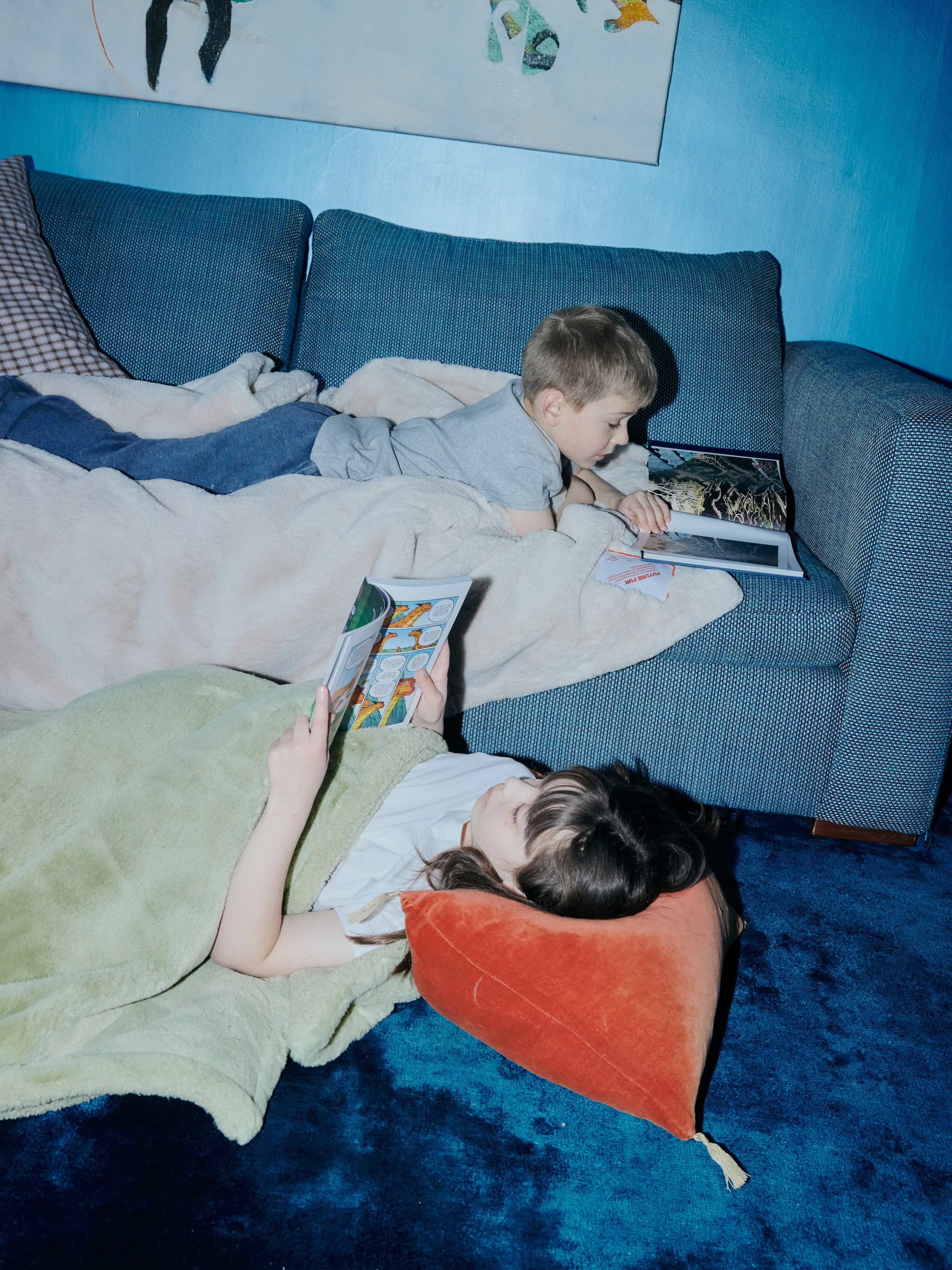Two children reading books while relaxing on a blue sofa and carpeted floor in a living room
