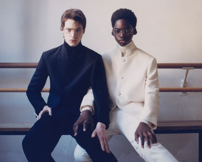 Two people, one with light skin and one with dark skin, wearing jackets and eyeglasses, are sitting indoors in a fashion editorial.