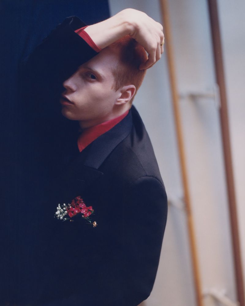 Portrait of red-haired male in dark suit with floral detail against two-tone backdrop in a studio with soft lighting and shallow depth of field.