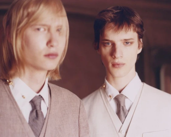 Two young men in formal attire, one with blond hair and the other with brown hair, pose in an indoor setting. Fashion shoot.