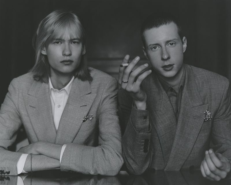 Monochrome portrait of two men in matching suits, one holding a cigarette, posing indoors with a neutral expression, formal attire.