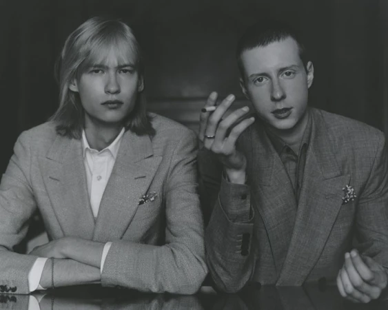 Monochrome portrait of two men in matching suits, one holding a cigarette, posing indoors with a neutral expression, formal attire.