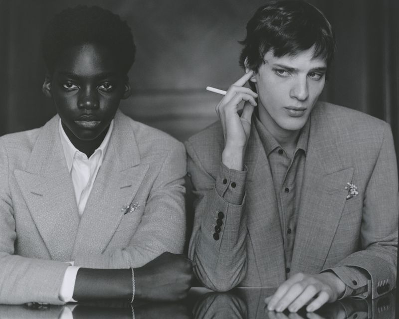 Two fashion models pose indoors. One holds a cigarette. They wear grey suits with collared shirts in a medium shot.