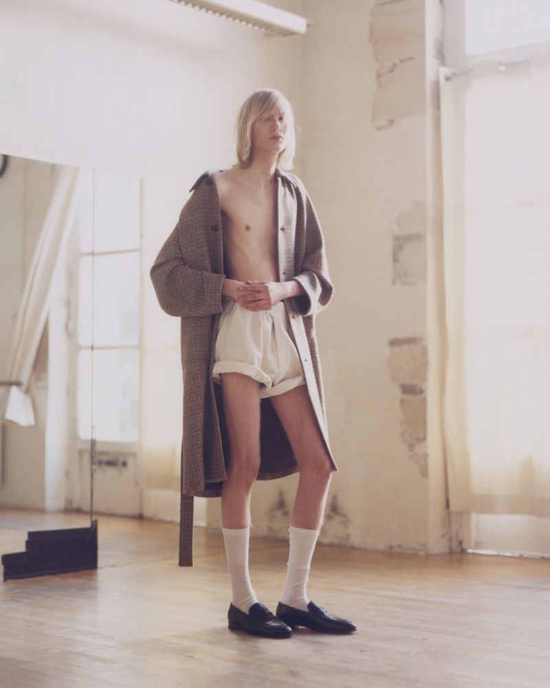 Male fashion model poses in an airy photo studio, wearing a wool coat, white shorts, socks, and loafers. Soft, natural light.