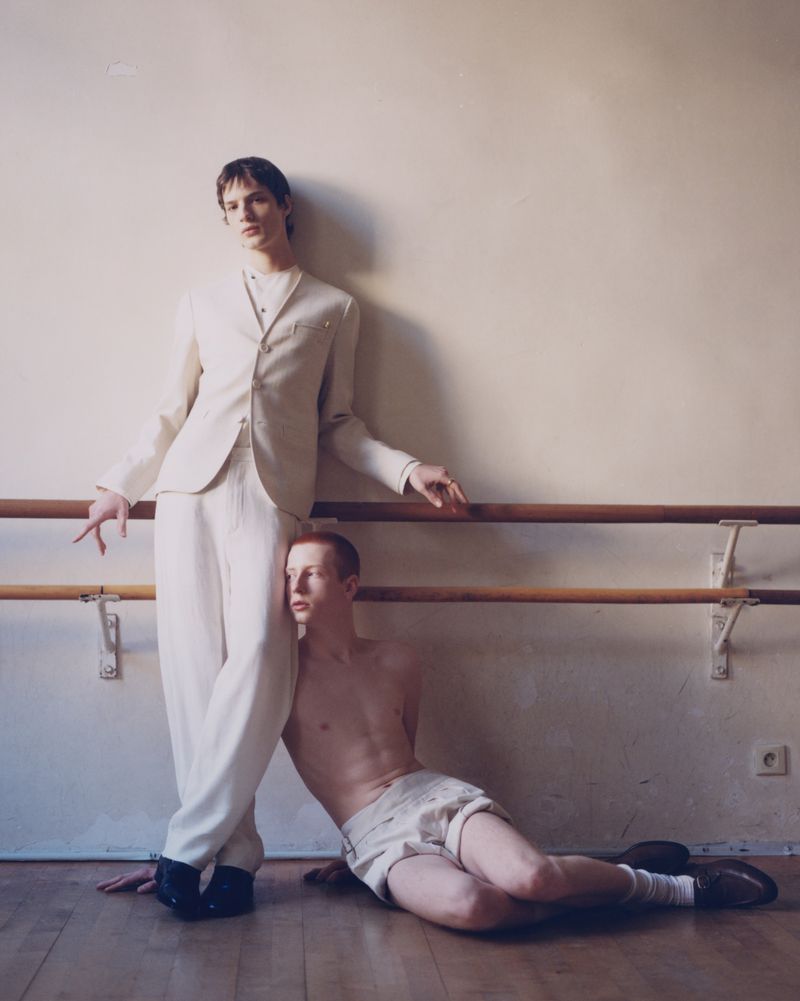 Two young men pose in a dance studio with a barre. One is dressed in a white suit, while the other is shirtless in shorts.