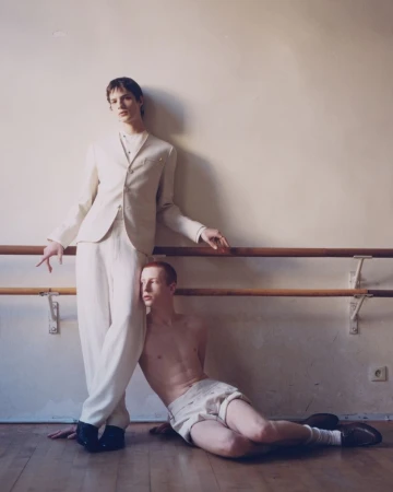 Two young men pose in a dance studio with a barre. One is dressed in a white suit, while the other is shirtless in shorts.