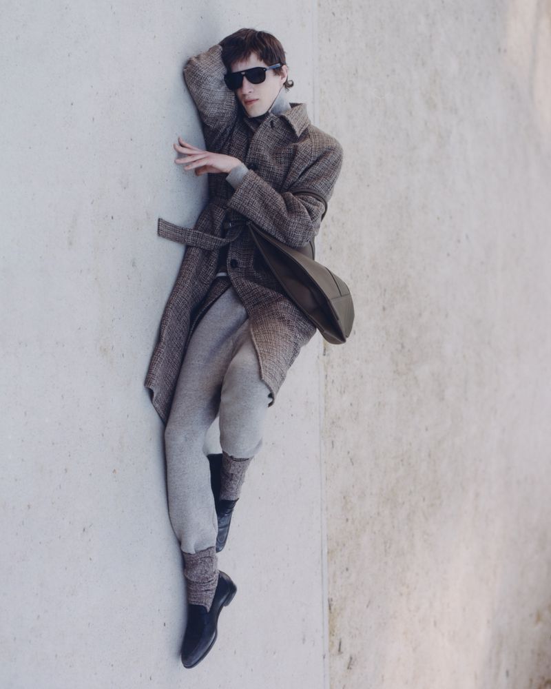 Fashion portrait of a man in a coat and sunglasses, reclining against a wall in a studio setting with neutral colors and soft lighting.