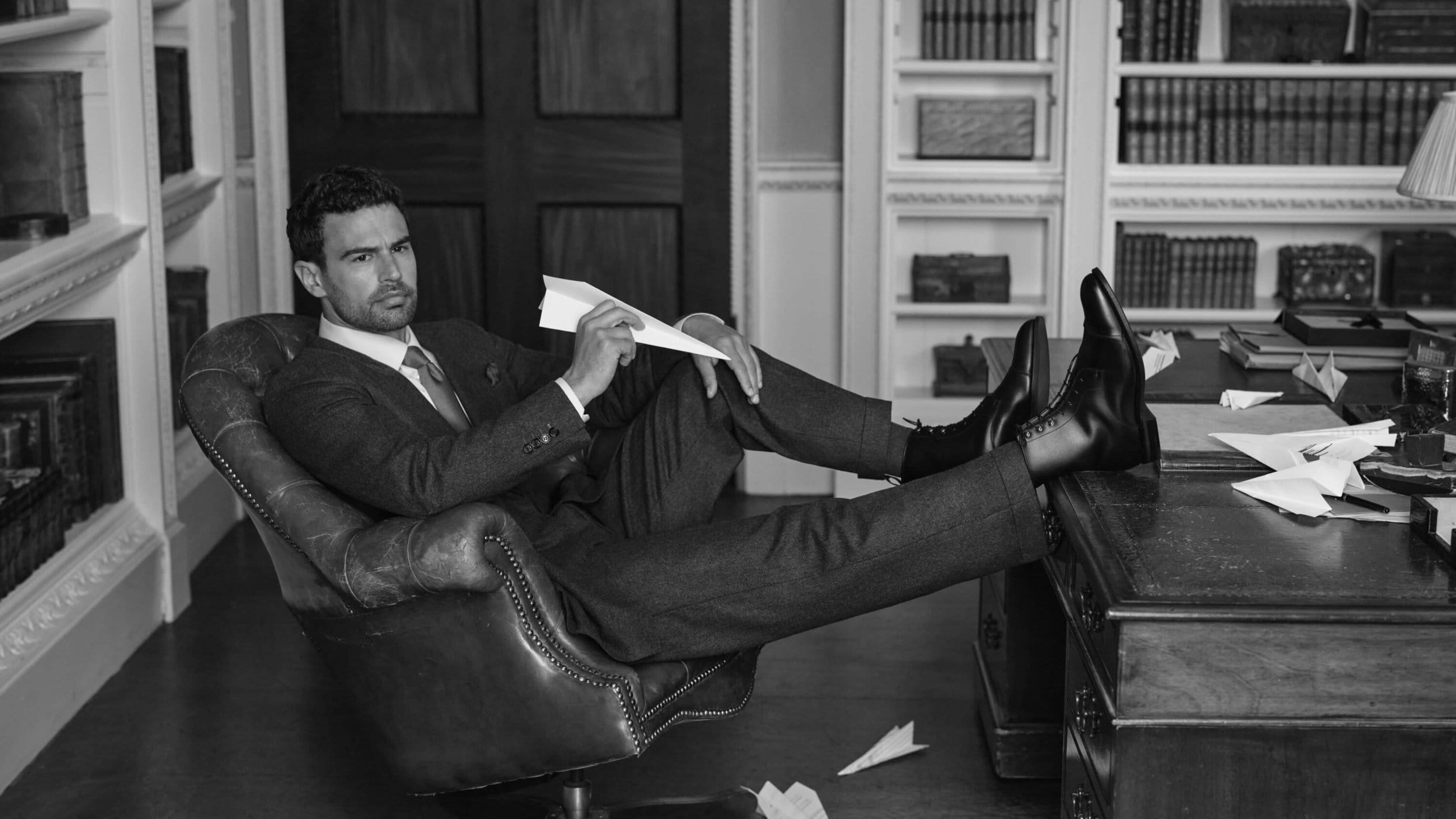 Theo James in a suit holding a paper airplane with his feet up on a desk in a library for Church's Fall 2025 campaign.