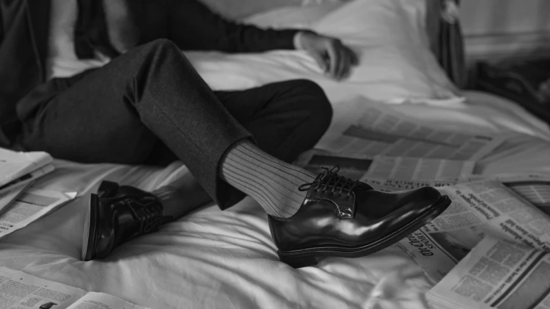 Man wearing leather dress shoes and ribbed socks resting on a white bed with scattered newspapers