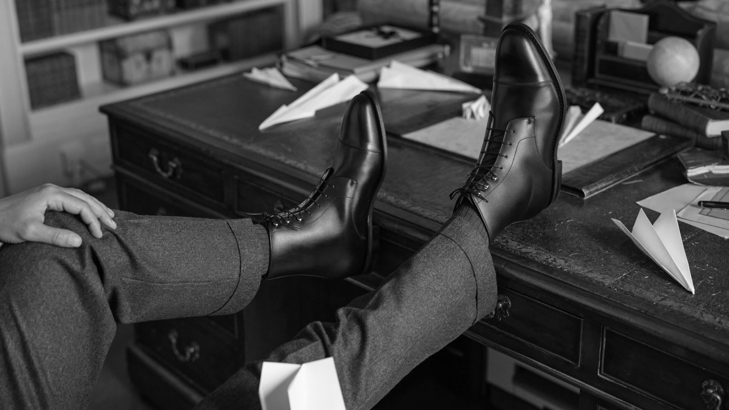Close-up of polished black leather dress boots worn with dark trousers, resting on a vintage wooden desk.