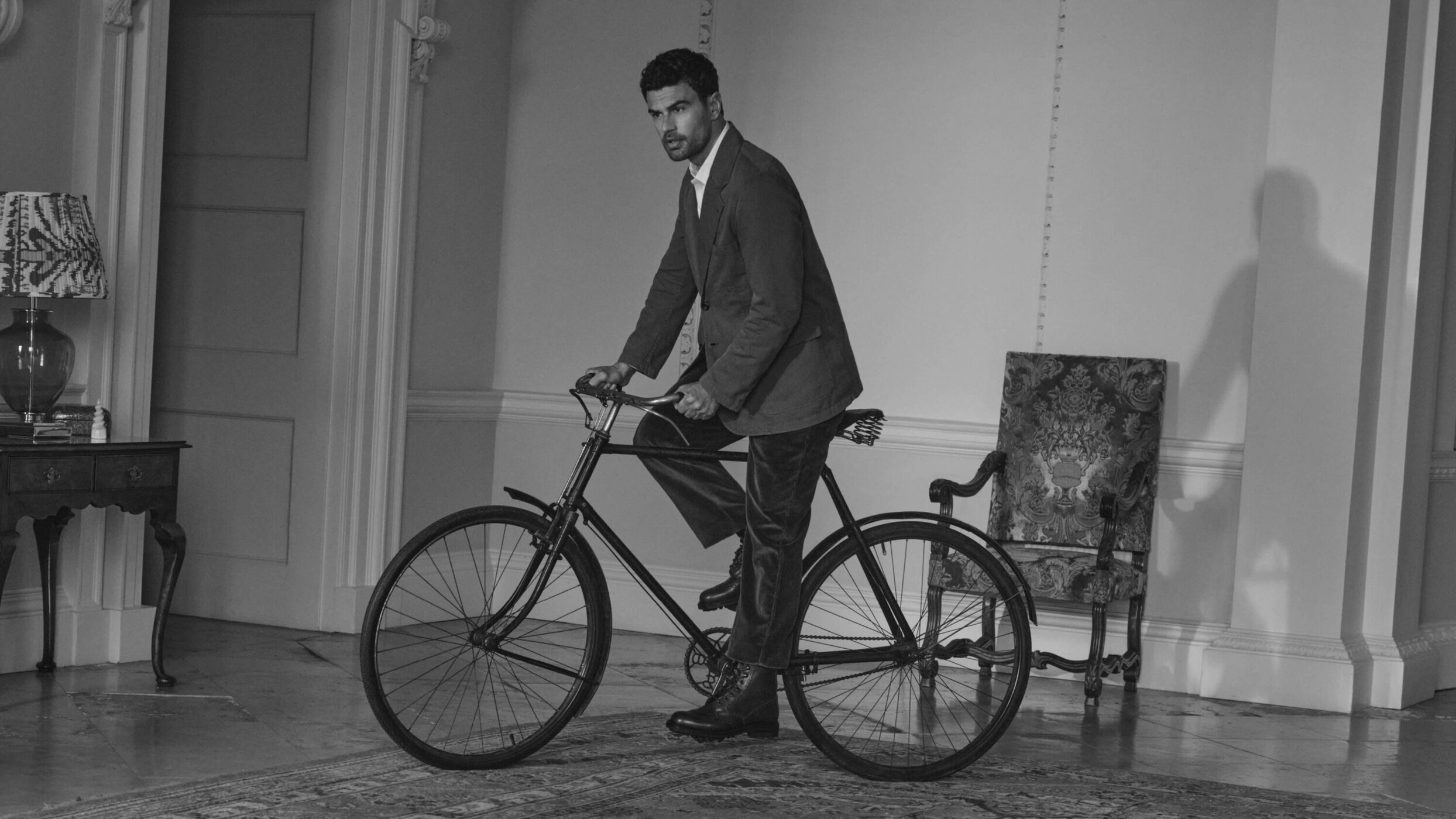A man in a suit jacket posing on a vintage bicycle inside a classic room with patterned chairs and classic architecture.